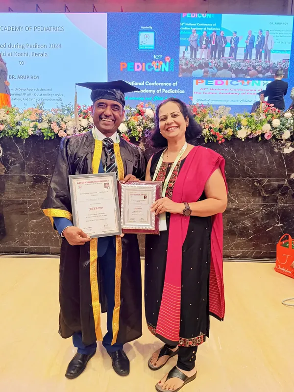 Dr. Ramakant Patil in academic regalia holding certificates, standing with Dr. Sadhana Patil in front of a conference stage.